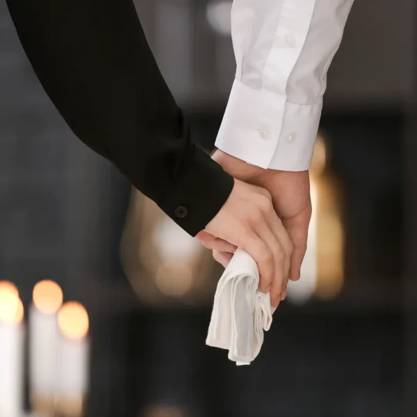 Zwei Menschen in dunkler Kleidung halten sich still an den Händen, eine Person hält ein weißes Tuch – symbolisches Bild für Abschied, Würde und Vorsorge am Lebensende.