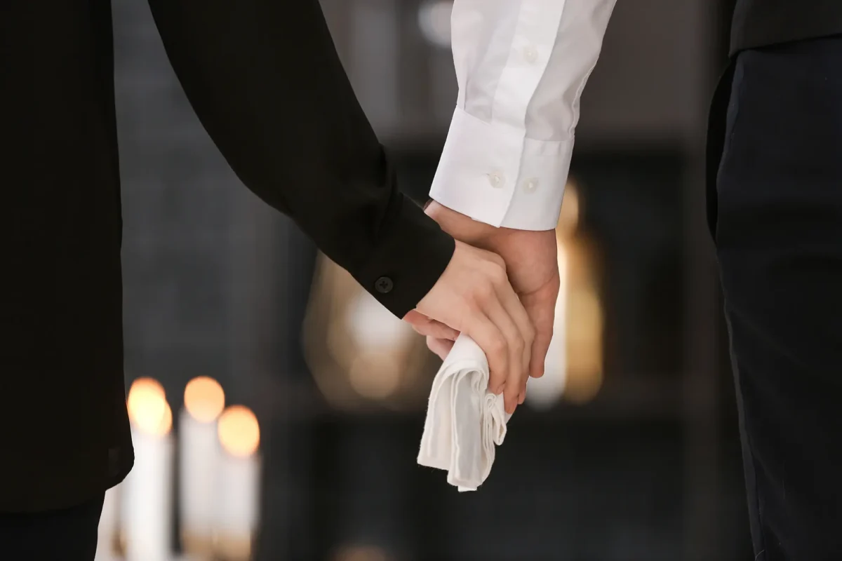 Zwei Menschen in dunkler Kleidung halten sich still an den Händen, eine Person hält ein weißes Tuch – symbolisches Bild für Abschied, Würde und Vorsorge am Lebensende.