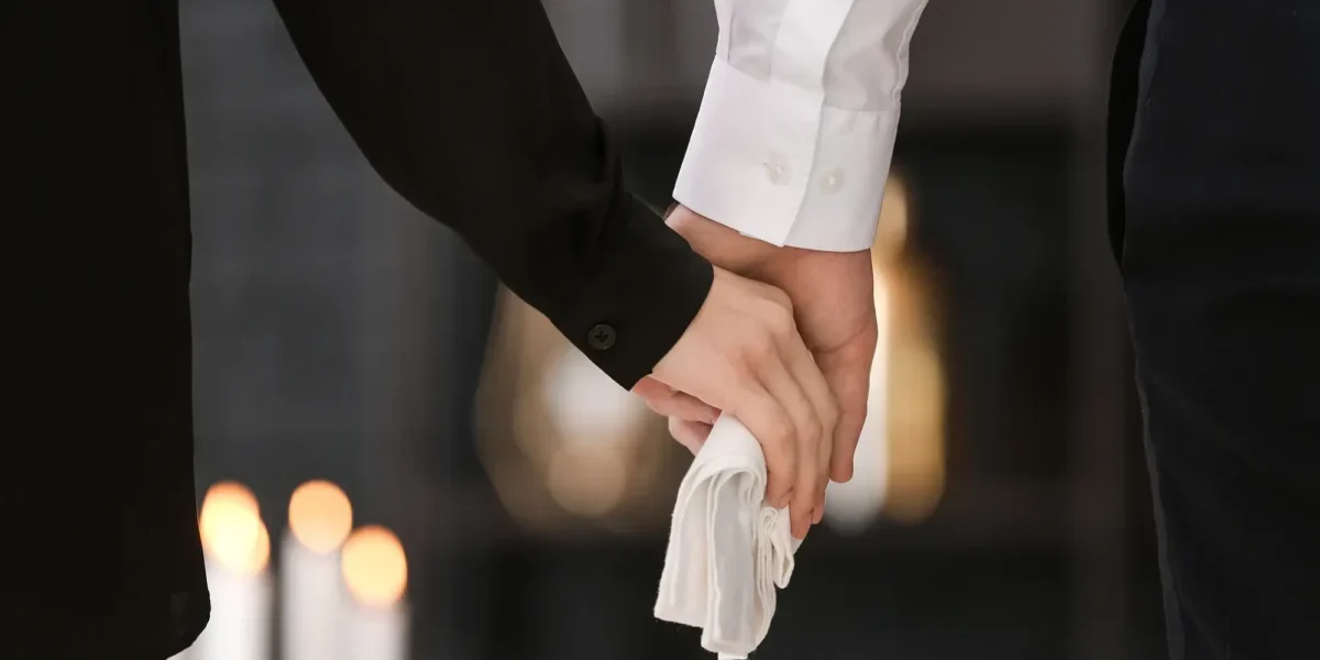 Zwei Menschen in dunkler Kleidung halten sich still an den Händen, eine Person hält ein weißes Tuch – symbolisches Bild für Abschied, Würde und Vorsorge am Lebensende.